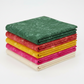Expressions Batiks Hand-Dyes Evergreen fabric by Riley Blake Designs on top of a neat folded stack of coordinating Expressions Batiks Hand-Dyes tonal batik fabrics in warm red, curry yellow, hot pink, and cream – rich deep forest green hand-dyed batik with organic mottled texture, 100% cotton
