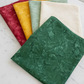 Expressions Batiks Hand-Dyes Evergreen fabric by Riley Blake Designs folded on a white marble surface with coordinating tonal batik swatches in warm red, golden yellow, soft sage, and cream nearby – rich deep forest green hand-dyed batik with organic mottled texture, a versatile tonal blender perfect for nature-inspired and jewel-tone quilts, 100% cotton batik