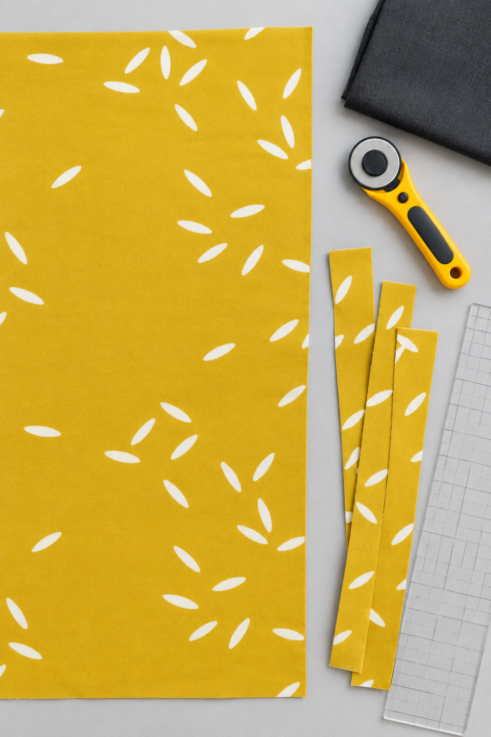 Filigree Rice Saffron fabric by Zen Chic for Moda Fabrics laid flat with cut strips, a rotary cutter, a clear quilting ruler, and a folded charcoal coordinating fabric. Available at IraRobi Quilts.