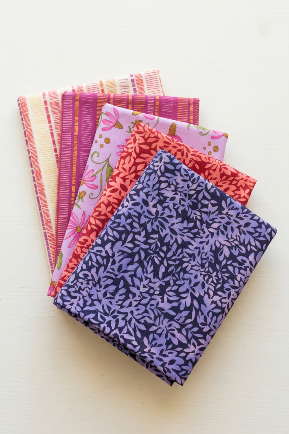 Five folded Superbloom fabrics by Robin Pickens for Moda Fabrics arranged in a fan, featuring Leafy Indigo, Leafy Geranium, Daisyish Freesia, and Orchid and Cream Scribble Stripes.