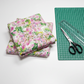 Folded Decoupage Pink Lilac quilting fabric beside a green cutting mat, clear ruler, and fabric scissors.