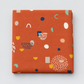 Frisky Catch Me Cheeky fabric by Zen Chic for Moda Fabrics, neatly folded on a light grey background, showing navy, mint, mustard, cream, and blush abstract shapes and dotted clusters on a rust-orange ground.