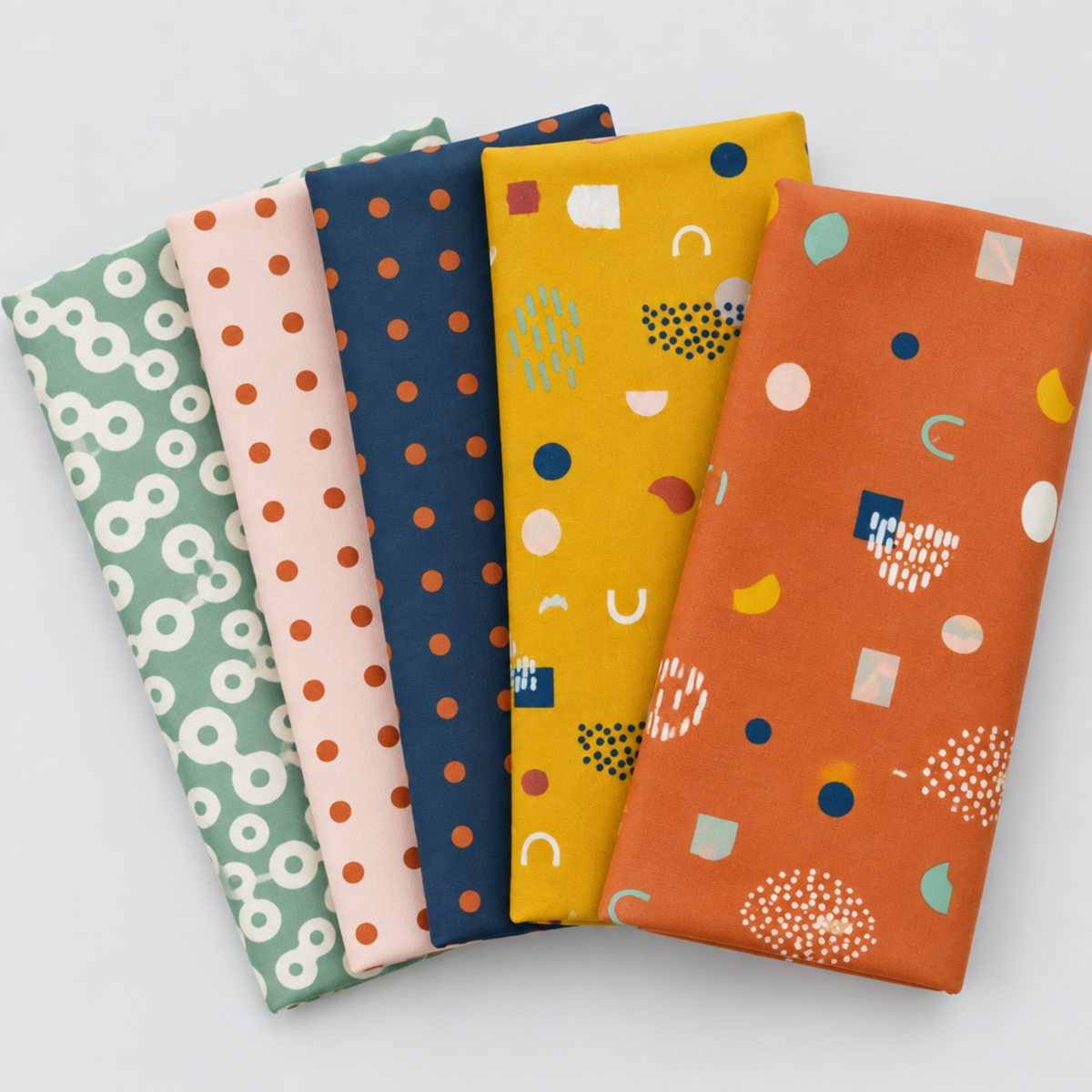Frisky fabric collection by Zen Chic for Moda Fabrics – fanned stack of Floating Chill, In A Row Sweetie, In A Row Moody, Catch Me Sunny, and Catch Me Cheeky coordinating prints in sage, blush, navy, mustard, and rust on a light grey background. All available at IraRobi Qui