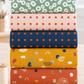 Coordinating Frisky fabrics by Zen Chic for Moda Fabrics stacked on a sewing table – Floating Chill sage rings, In A Row Sweetie blush dots, In A Row Moody navy dots, Catch Me Sunny mustard shapes, and Catch Me Cheeky rust shapes, with thread and gold scissors. All available at IraRobi Quilts.