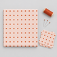 Frisky In A Row Sweetie fabric by Zen Chic for Moda Fabrics folded beside a stack of cut squares, a spool of rust-orange thread, and pins on a light grey background.