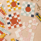 Garden Afternoons quilt blocks in progress with Frisky fabrics by Zen Chic for Moda Fabrics – finished ring blocks with rust-orange centers and one block laid out in loose squares, beside remnant scraps, folded fabrics, and sewing tools on a wooden table. Quilts shown for inspiration feature the Garden Afternoons Quilt Pattern by Zen Chic — all available at IraRobi Quilts.