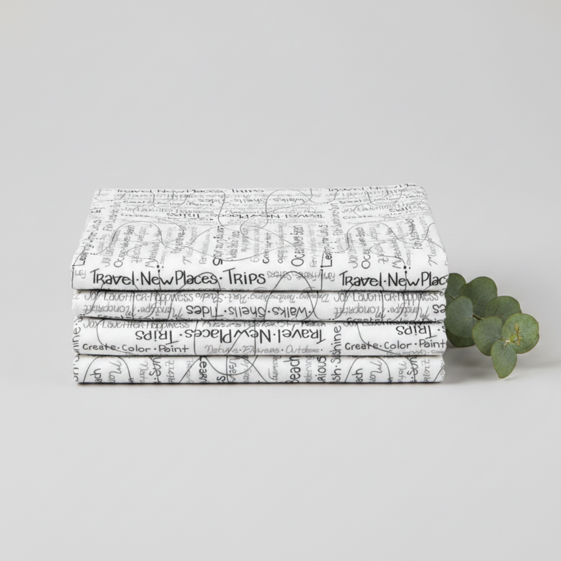 Writing on the Wall Going Places White quilting cotton by Sew Kind of Wonderful for FreeSpirit Fabrics -- three folded yards stacked on a light grey surface with a eucalyptus sprig, crisp white ground with handwritten black travel and lifestyle words including Travel, Joy, Ocean, Waves, Beach, Sunshine, Create, and Nature