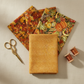 Autumn Celebration Gold Berries quilting cotton — folded fat quarter as front hero with coordinating Autumn Celebration autumn leaf and Multi Vegetables harvest prints fanned behind. Brass scissors and amber thread spools on cream linen. Coordinating fabrics and quilt patterns available at IraRobi Quilts.