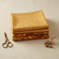 Autumn Celebration Gold Berries quilting cotton — folded as top hero on a neat stack with coordinating autumn leaf and Multi Vegetables harvest prints beneath. Brass scissors and tied acorn bundle on cream linen. Coordinating fabrics and quilt patterns available at IraRobi Quilts.