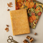 Autumn Celebration Gold Berries quilting cotton — folded fat quarter as front hero with coordinating Autumn Celebration fabrics fanned behind: rich autumn leaf print and Multi Vegetables harvest print with pumpkins and gourds. Brass scissors and amber thread spools on cream linen. Coordinating fabrics and quilt patterns available at IraRobi Quilts.