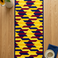 Golden Weave quilted table runner centered on a light wood tabletop beside a pitcher of sunflowers and a cream cup and saucer on a folded navy napkin.