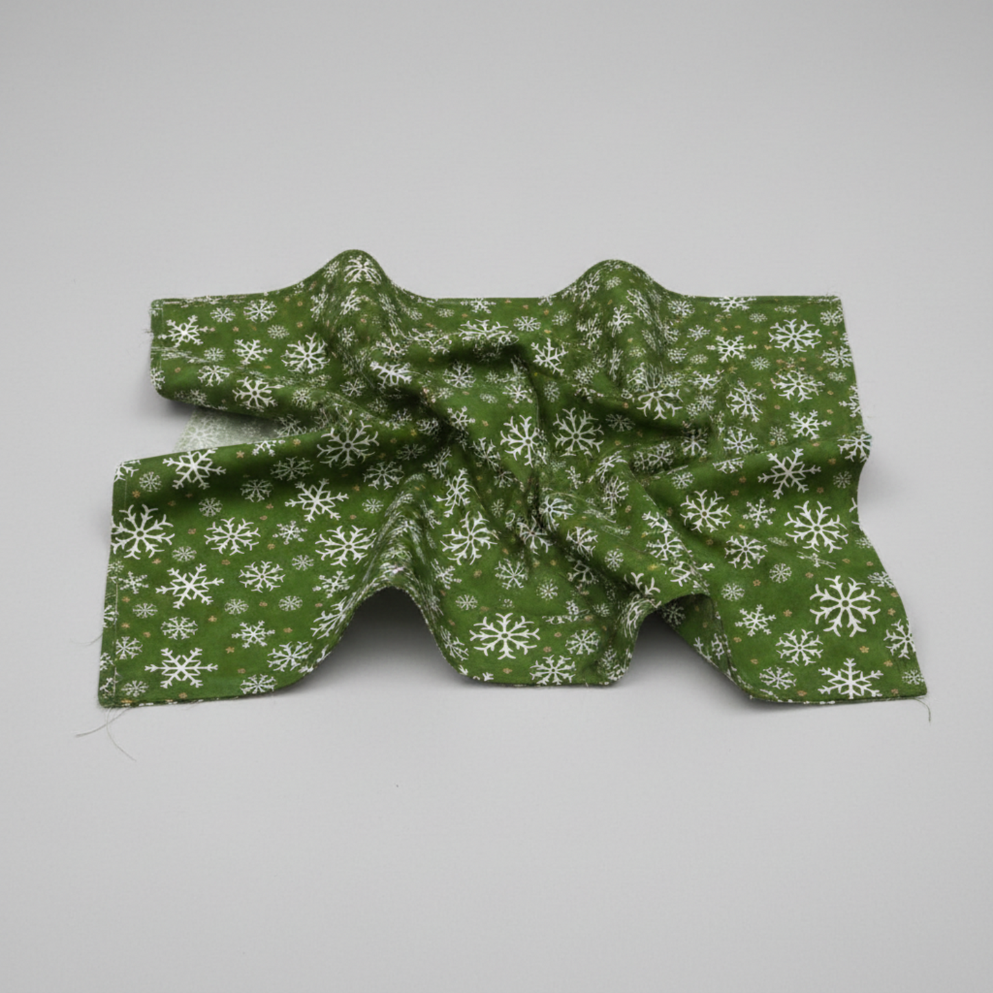 Winter Solstice II Green Snowflakes Metallic quilting cotton by Jason Yenter for In The Beginning Fabrics — fabric draped over a grey surface showing natural folds and raw cut edge, rich deep forest green with tossed white snowflakes and gold metallic star dots. Quilt blocks shown for inspiration. Coordinating fabrics and quilt patterns are available at IraRobi Quilts.