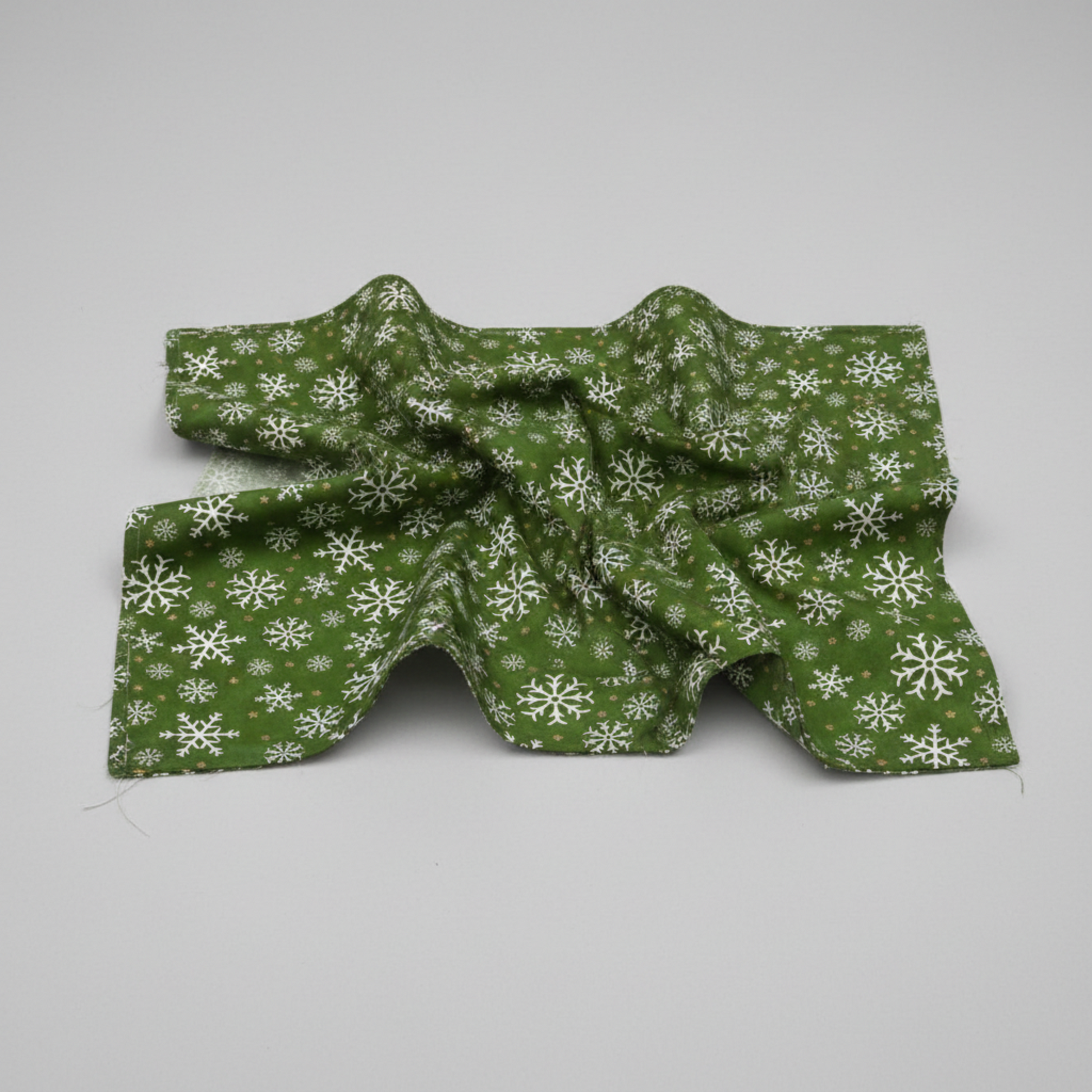 Winter Solstice II Green Snowflakes Metallic quilting cotton by Jason Yenter for In The Beginning Fabrics — fabric draped over a grey surface showing natural folds and raw cut edge, rich deep forest green with tossed white snowflakes and gold metallic star dots. Quilt blocks shown for inspiration. Coordinating fabrics and quilt patterns are available at IraRobi Quilts.