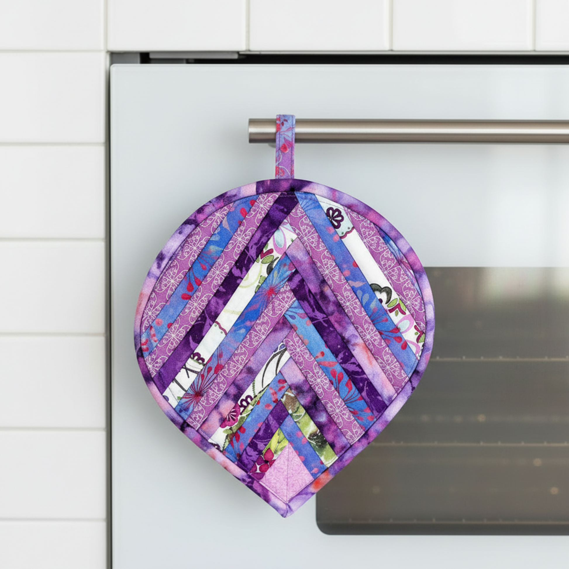Leaf-Shaped Quilted Hot Pad – Purple Garden hanging upside down on a stainless steel oven handle, pointed tip down, loop hooked over the bar, showing herringbone chevron strip piecing in purple, lavender, blue, pink, magenta, and floral botanical prints – handmade by IraRobi Quilts