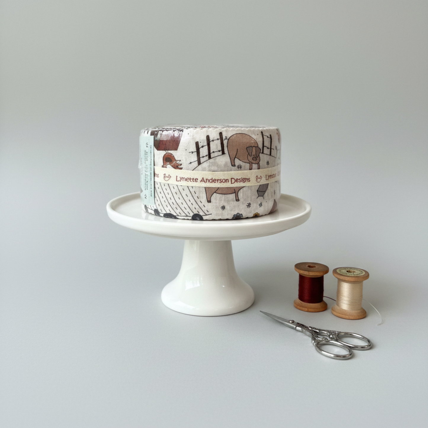 High Meadow Farm Jelly Roll by Lynette Anderson for RJR Fabrics on a white cake stand with embroidery scissors and wooden thread spools