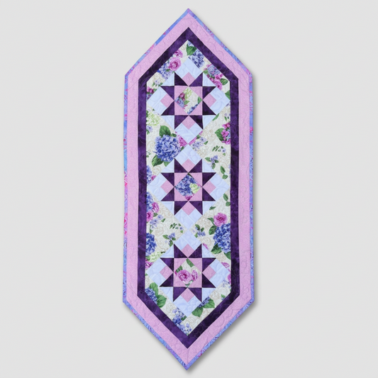 Bread and Butter Hydrangea Bloom quilted table runner with three purple star blocks, hydrangea and rose prints, and lavender borders on light gray.