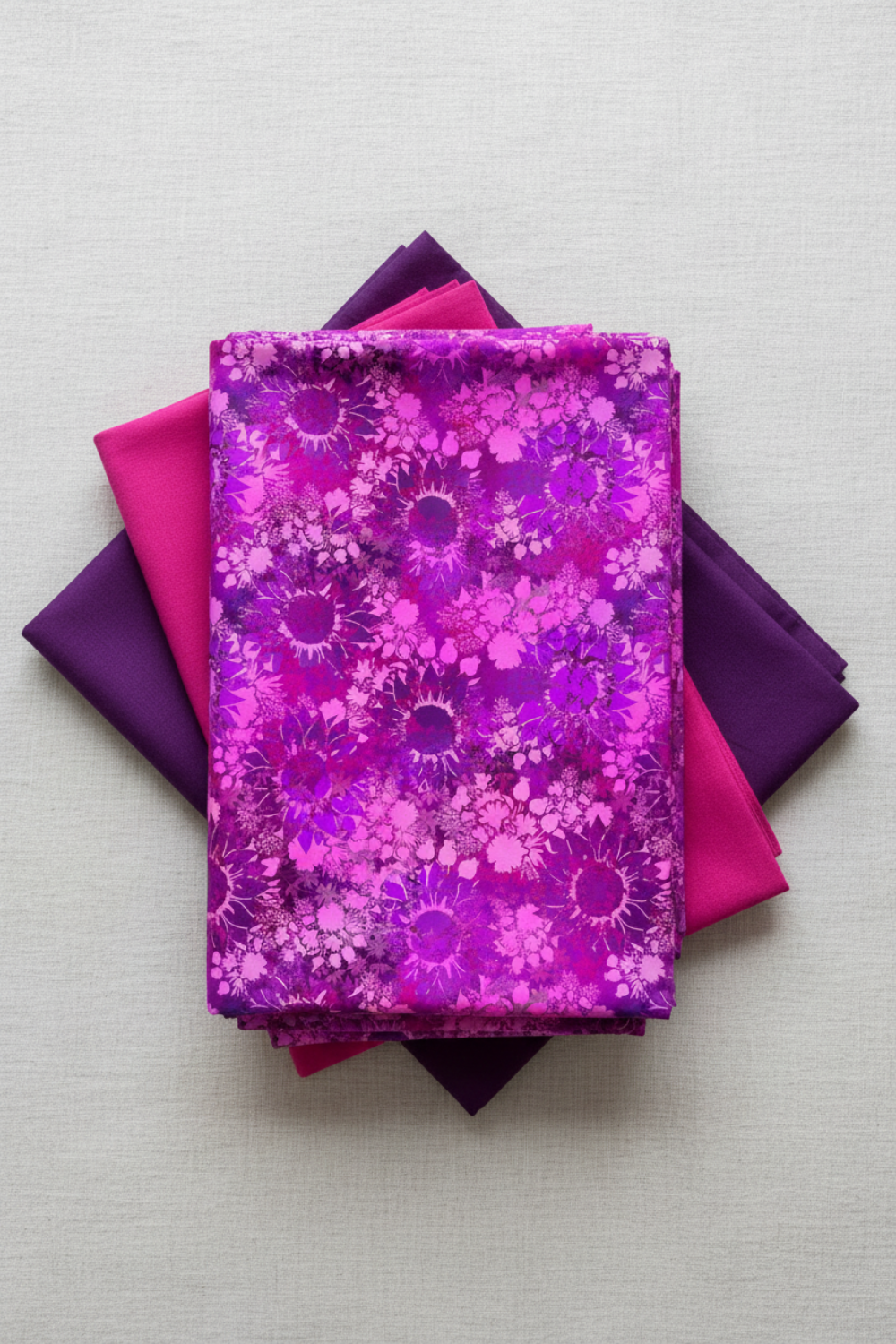 I Dream In Color Sunflower Magenta fabric by Jason Yenter for In The Beginning Fabrics folded on a light linen surface with coordinating jewel-toned pink and purple fabrics – dense allover tonal shadow floral with silhouetted sunflowers and daisy clusters in magenta, hot pink, fuchsia, and deep purple watercolor wash, 100% cotton, 3 yard cut