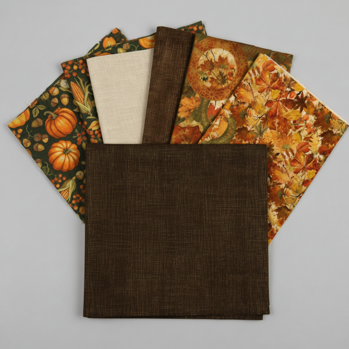 Interwoven Bark Fabric by Jason Yenter for In The Beginning Fabrics fanned with coordinating autumn fabrics – olive crosshatch, autumn medallion leaves print, harvest pumpkins and gourds print, and packed rust and orange leaves print