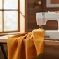 Interwoven Butterscotch Fabric by Jason Yenter for In The Beginning Fabrics draped in generous folds next to a white sewing machine on a wooden sewing table — warm golden amber crosshatch tonal with linen-like texture depth. 100% cotton quilting cotton.