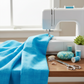 Interwoven Deep Sky Fabric by Jason Yenter for In The Beginning Fabrics draped in soft folds next to a white sewing machine — vivid bright sky blue crosshatch tonal with shells, teal thread spools, and a small potted plant alongside. 100% cotton quilting cotton, 43–44" wide.