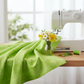 Interwoven Lime Fabric by Jason Yenter for In The Beginning Fabrics draped in generous folds next to a white sewing machine on a wooden sewing table — vivid lime green crosshatch tonal with fresh spring flowers and green leaves as styling props. 100% cotton quilting cotton.