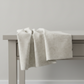 Interwoven Taupe Fabric by Jason Yenter for In The Beginning Fabrics — soft cream and taupe linen-look crosshatch blender quilting cotton draped over a grey table edge in a clean studio setting, raw cut edges visible, 100% cotton, 43–44" wide
