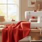Interwoven Tomato Fabric by Jason Yenter for In The Beginning Fabrics draped in generous folds next to a white sewing machine on a wooden sewing table — vivid fire-engine red crosshatch tonal with linen-like texture depth. 100% cotton quilting cotton.