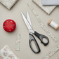 KAI N5360 9in Pinking Shears with black and gray ergonomic handles on a linen surface, surrounded by folded floral and solid quilting fabric swatches, pinked zigzag-edge fabric strips, a red tomato pin cushion with colorful pins, and a spool of white thread