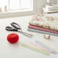 AI N5360 9in Pinking Shears with black and gray ergonomic handles on a white sewing table beside a red tomato pin cushion, a stack of folded floral, gingham, and ditsy print quilting fabrics, a wooden thread spool, and fabric strips with clean zigzag pinked edges in a bright natural-light sewing room