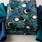 La Luna Blue Silver Moon Collage fabric by Jason Yenter for In The Beginning Fabrics folded in a stack on a light linen surface with coordinating deep teal and navy fabrics – dramatic moon phase medallions with silver radiating lines, crescent moons, and watercolor botanicals in teal, emerald, and cobalt on deep black, perfect for celestial and night-themed quilt projects, 100% cotton, 3 yard cut