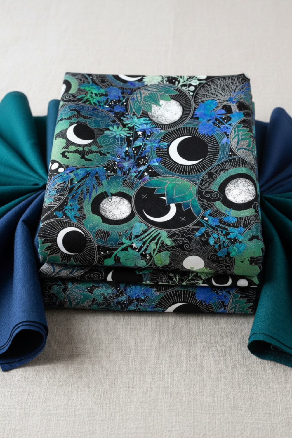 La Luna Blue Silver Moon Collage fabric by Jason Yenter for In The Beginning Fabrics folded in a stack on a light linen surface with coordinating deep teal and navy fabrics – dramatic moon phase medallions with silver radiating lines, crescent moons, and watercolor botanicals in teal, emerald, and cobalt on deep black, perfect for celestial and night-themed quilt projects, 100% cotton, 3 yard cut