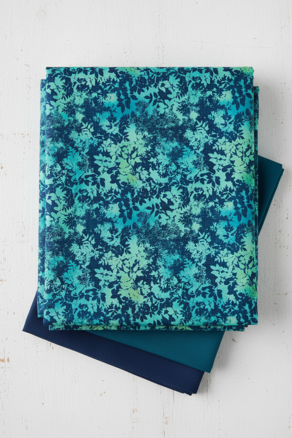 La Luna Teal Leaves fabric by Jason Yenter for In The Beginning Fabrics folded in a stack on a light linen surface with coordinating deep teal and navy fabrics – dense allover tonal leaf silhouette print with fern fronds and botanical sprigs in aqua, teal, chartreuse, and navy on rich cobalt watercolor wash, perfect for night garden quilt projects, 100% cotton, 3 yard cut