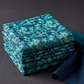 Luna Teal Leaves fabric by Jason Yenter for In The Beginning Fabrics folded in a stack on a dark linen surface with coordinating deep teal blender – dense allover tonal leaf silhouette print with fern fronds and botanical sprigs in aqua, teal, chartreuse, and navy on rich cobalt watercolor wash, perfect for night garden quilt projects, 100% cotton, 3 yard cut