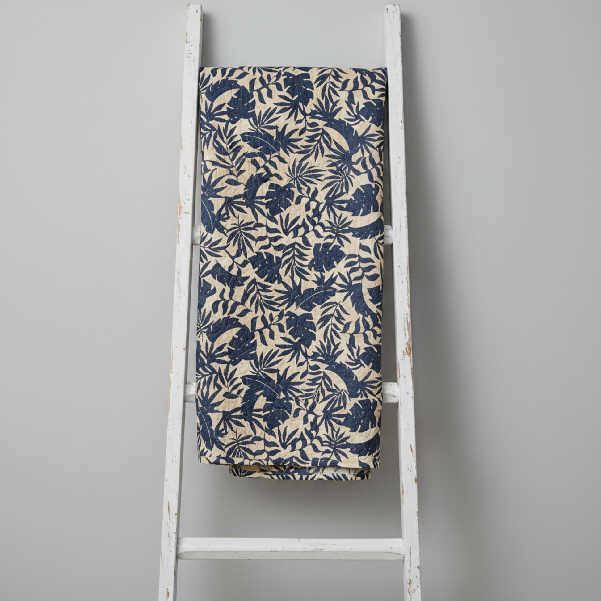 Breezeway Textured Palms Blue/Cream quilting cotton by Maywood Studio — fabric draped over a weathered white wooden ladder showing natural drape and bold navy-blue tropical palm and monstera silhouette print