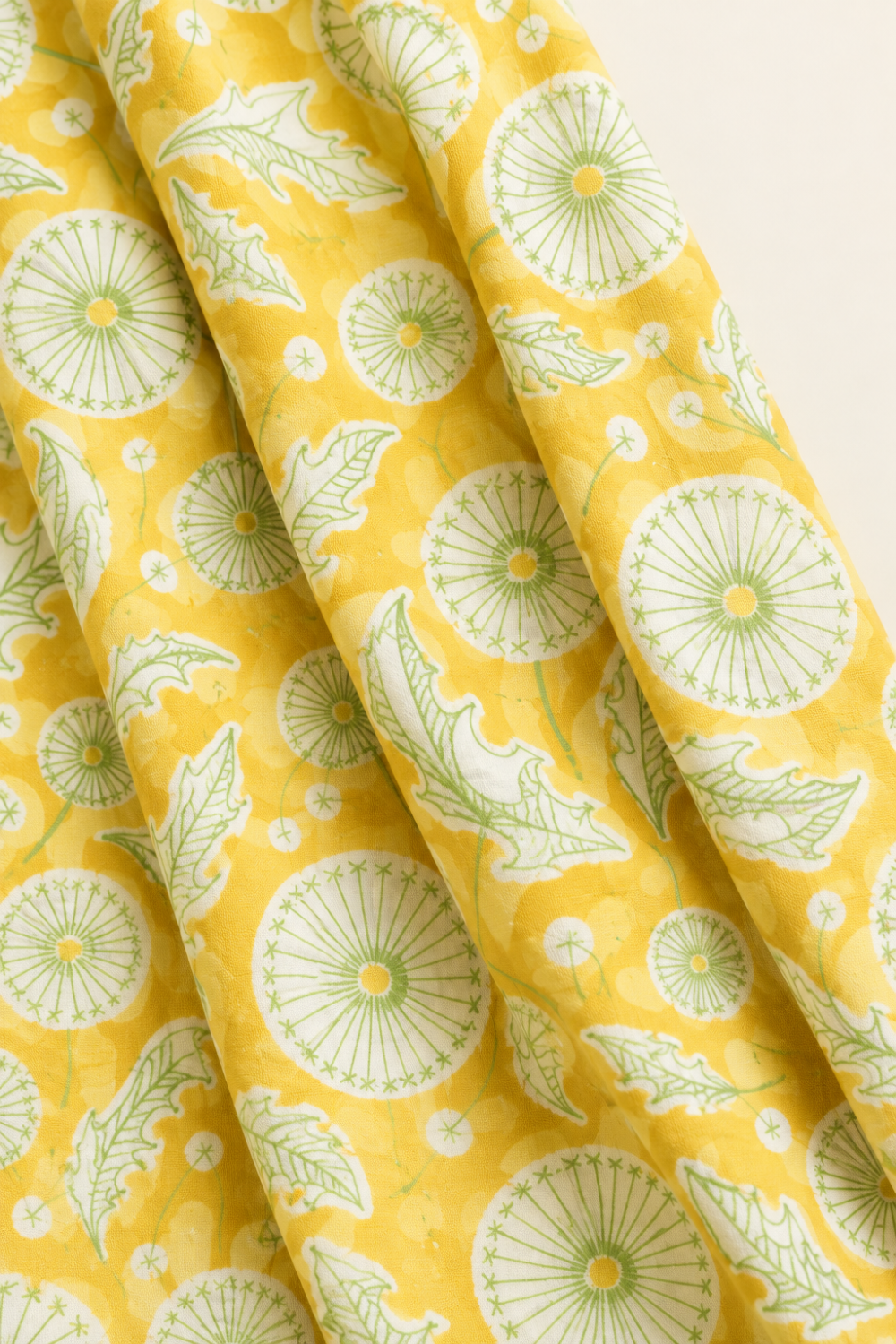 Folds of Dandi Duo Dandi Toile Maize fabric showing pale-green dandelion seed heads and leaves on a golden-yellow ground.