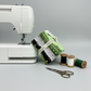 Maywood Studio Greenery Fat Quarter Bundle leaning against a white sewing machine with black, white and green thread spools and embroidery scissors on grey background