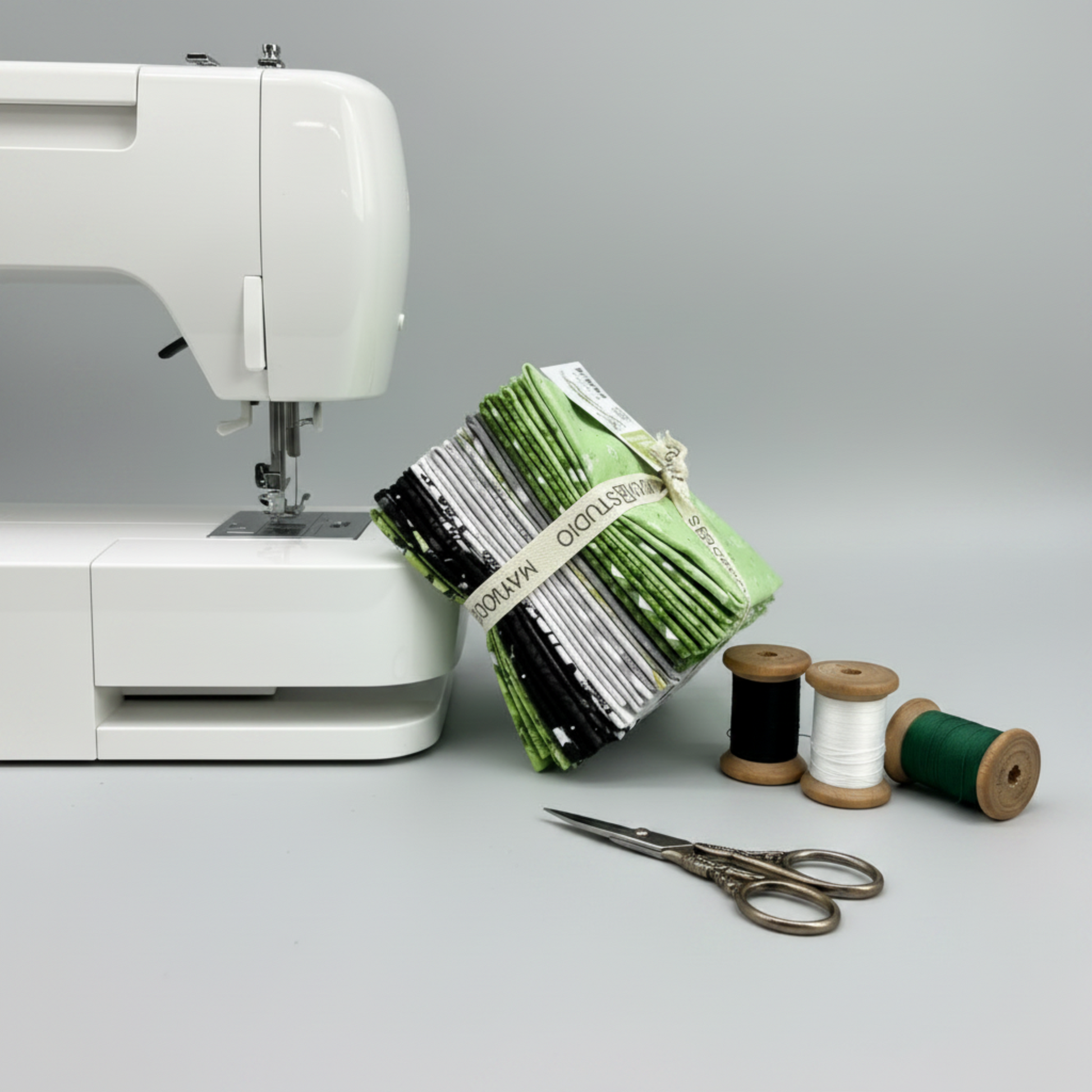 Maywood Studio Greenery Fat Quarter Bundle leaning against a white sewing machine with black, white and green thread spools and embroidery scissors on grey background