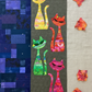ModCat quilt by Colourwerx work-in-progress featuring Expressions Batiks Hand-Dyes Bouquet fabric by Riley Blake Designs as the orange-red cat – vibrant multicolor hand-dyed batik with golden orange, hot pink, magenta, crimson, and lavender replacing the original orange batik, alongside yellow, green, and pink-red multicolor batik cats on a dark olive center panel with navy and purple scrappy patchwork background, loose cut Bouquet fabric pieces on a warm linen surface
