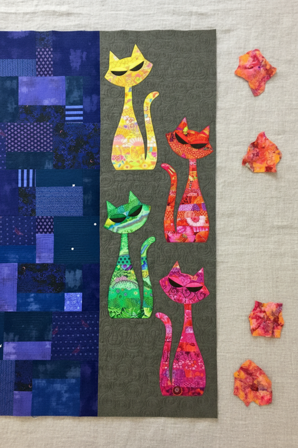 ModCat quilt by Colourwerx work-in-progress featuring Expressions Batiks Hand-Dyes Bouquet fabric by Riley Blake Designs as the orange-red cat – vibrant multicolor hand-dyed batik with golden orange, hot pink, magenta, crimson, and lavender replacing the original orange batik, alongside yellow, green, and pink-red multicolor batik cats on a dark olive center panel with navy and purple scrappy patchwork background, loose cut Bouquet fabric pieces on a warm linen surface