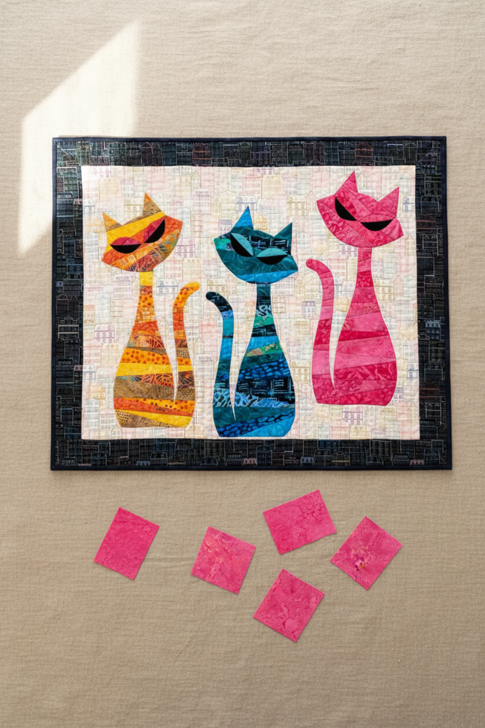 ModCat quilt by Colourwerx work-in-progress featuring Expressions Batiks Hand-Dyes Begonia fabric by Riley Blake Designs as the right cat – pure saturated hot pink hand-dyed batik with organic painterly texture replacing the original purple, alongside orange-gold and teal-blue striped tonal batik cats on a cream cityscape batik background with dark charcoal batik border, loose cut Begonia pink fabric pieces on a warm linen surface
