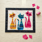 ModCat quilt by Colourwerx work-in-progress featuring Expressions Batiks Hand-Dyes Begonia fabric by Riley Blake Designs as the right cat – pure saturated hot pink hand-dyed batik with organic painterly texture, alongside orange-gold and teal-blue striped tonal batik cats on a cream cityscape batik background, with loose cut Begonia pink fabric pieces and pink thread spool on a warm linen surface