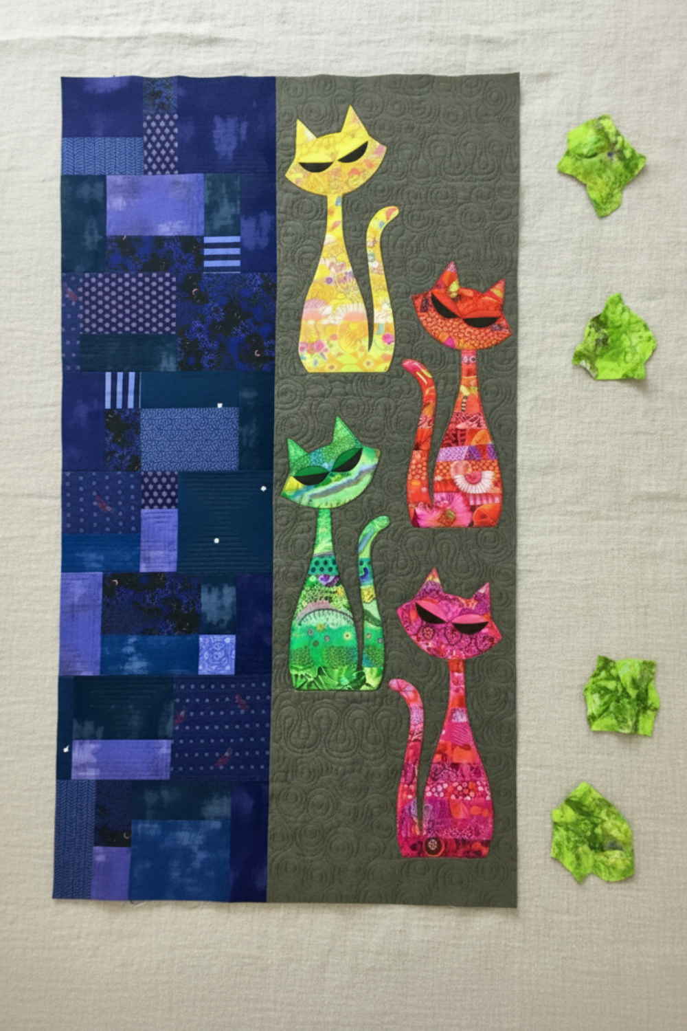 ModCat quilt wall hanging featuring Expressions Batiks Hand-Dyes Shamrock fabric by Riley Blake Designs as the green cat appliqué silhouette and crumpled fabric scraps on the right – bright electric lime-green hand-dyed batik with lively chartreuse yellow and soft teal-blue organic mottled overlays, with yellow, orange-red, and hot pink multicolor batik cat silhouettes on dark olive background, navy indigo patchwork panel, on natural linen. Quilt shown for inspiration features the ModCat Quilt Pattern by Co