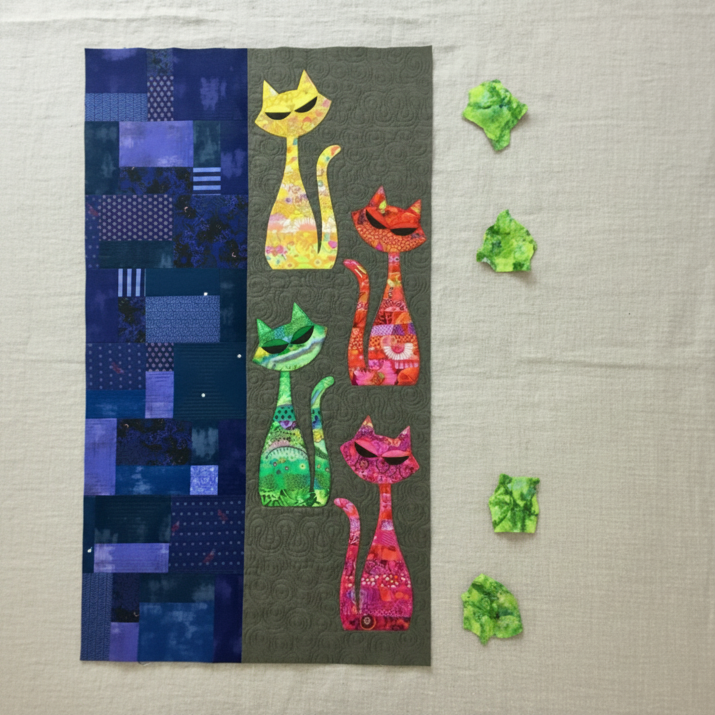ModCat quilt wall hanging featuring Expressions Batiks Hand-Dyes Shamrock fabric by Riley Blake Designs as the green cat appliqué silhouette and crumpled fabric scraps on the right – bright electric lime-green hand-dyed batik with lively chartreuse yellow and soft teal-blue organic mottled overlays, with yellow, orange-red, and hot pink multicolor batik cat silhouettes on dark olive background, navy indigo patchwork panel, on natural linen. Quilt shown for inspiration features the ModCat Quilt Pattern by Co