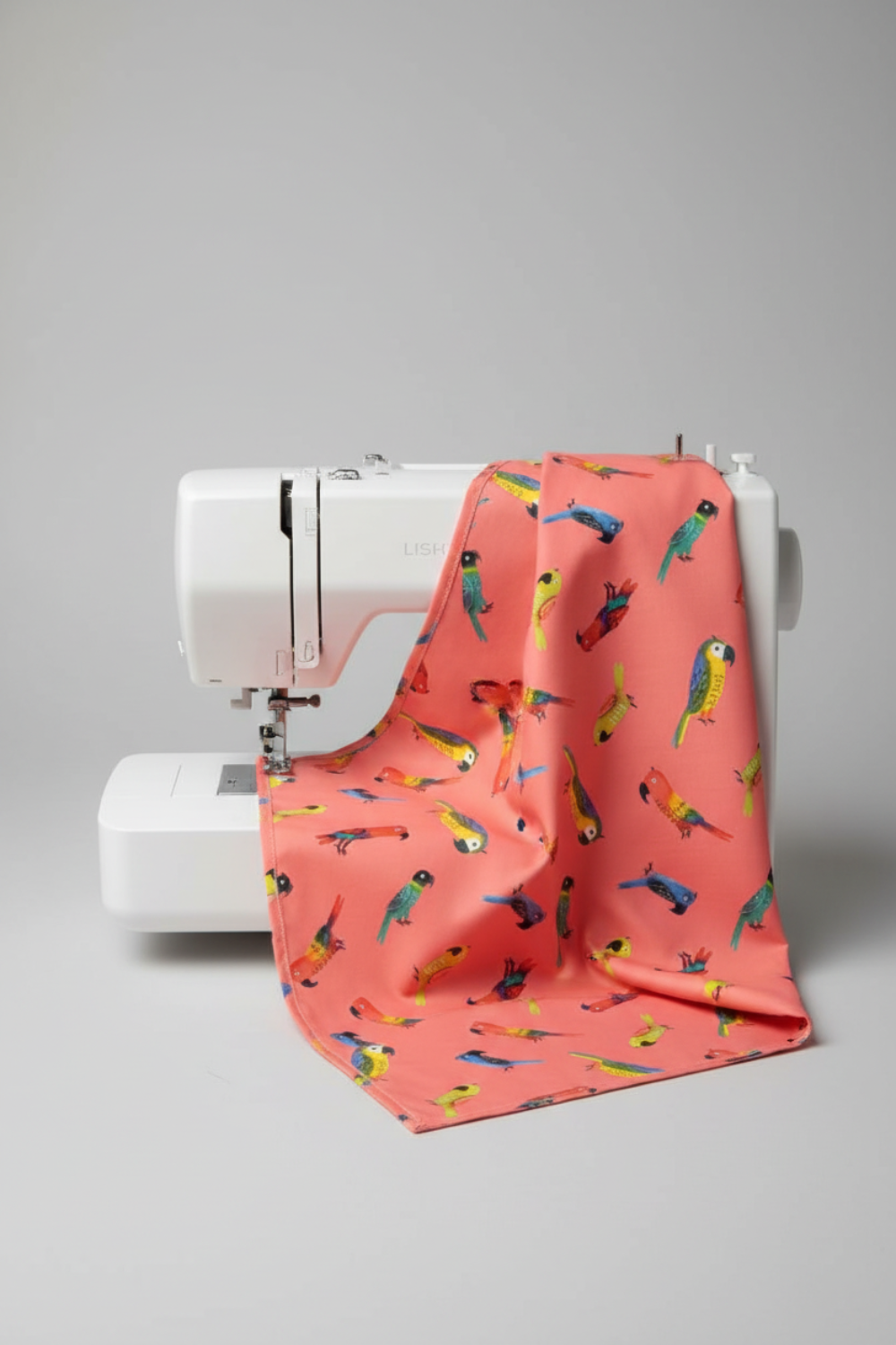 Coral sorbet pink quilting cotton with colorful tropical parrots draped over a modern white sewing machine — Safari Fun Jungle Birds Sorbet by Terry Runyan for Benartex