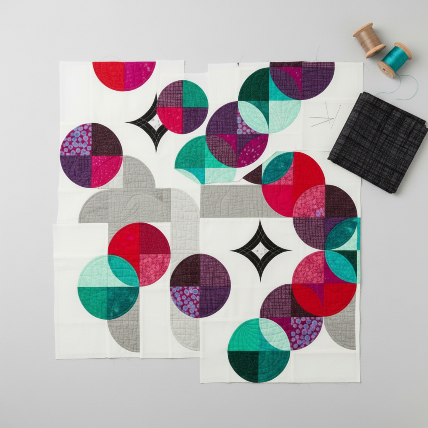 Interwoven Charcoal quilting cotton fabric cut into Moondance curved quilt pieces arranged on a light grey surface with a folded fabric swatch, straight pins, and thread spools – Interwoven Charcoal by Jason Yenter for In The Beginning Fabrics