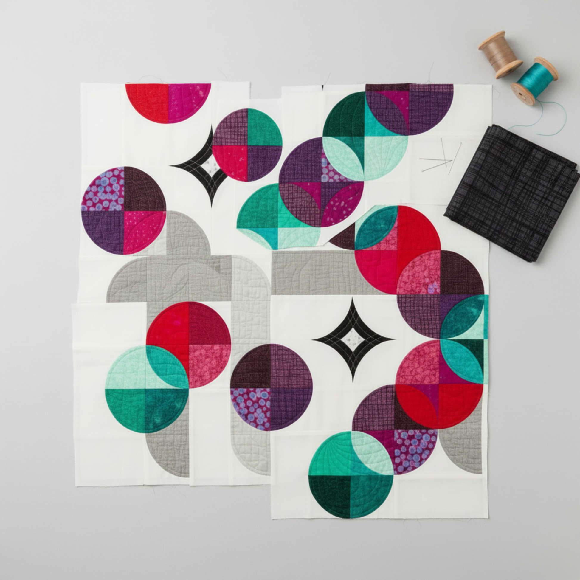 Interwoven Charcoal quilting cotton fabric cut into Moondance curved quilt pieces arranged on a light grey surface with a folded fabric swatch, straight pins, and thread spools – Interwoven Charcoal by Jason Yenter for In The Beginning Fabrics