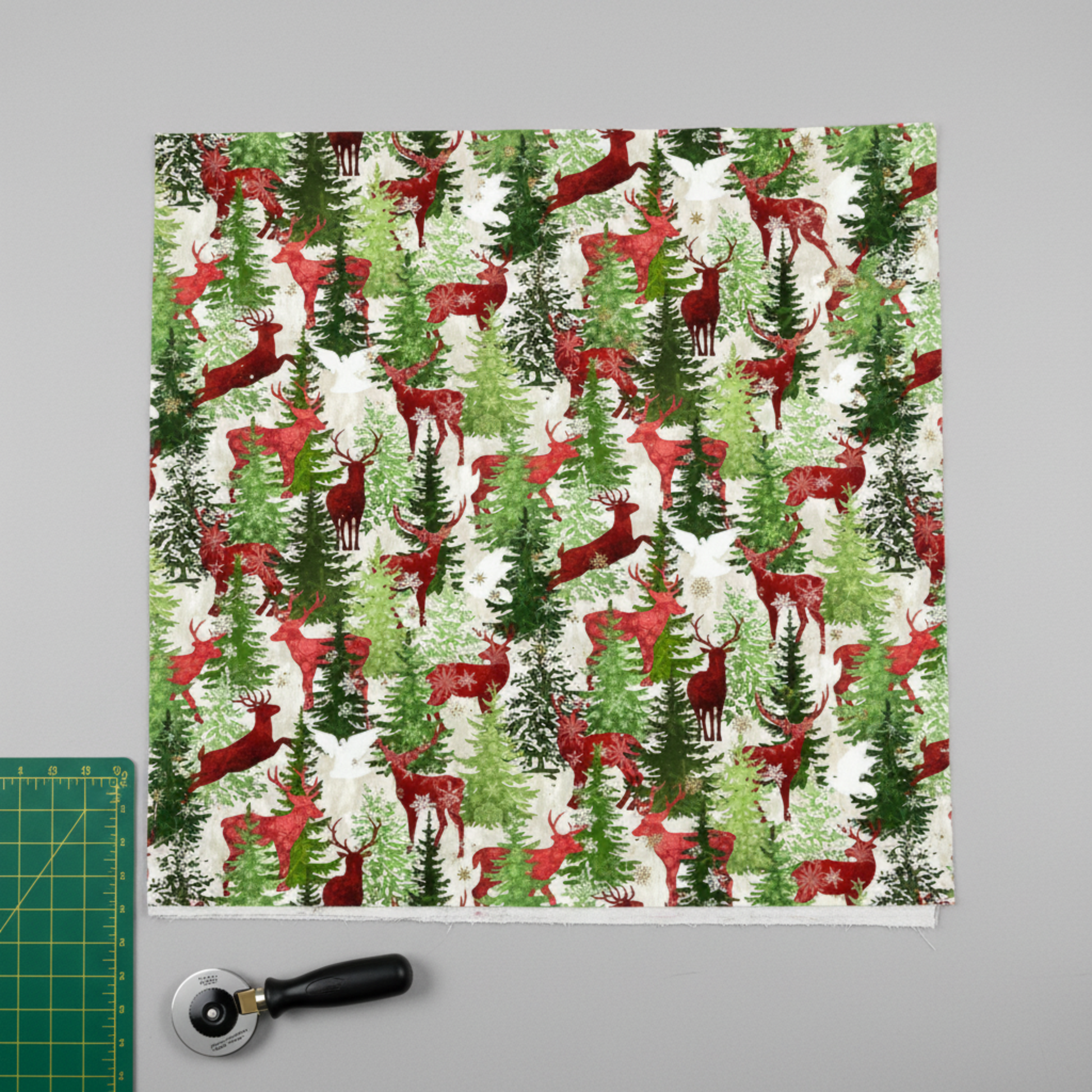 Winter Solstice II Multi Deer Metallic quilting cotton by Jason Yenter for In The Beginning Fabrics — fabric flat lay showing dramatic winter forest print with red deer among watercolor evergreen trees, snowflakes, and gold metallic star dots on cream, shown with cutting mat and rotary cutter. Quilt blocks shown for inspiration. Coordinating fabrics and quilt patterns are available at IraRobi Quilts.