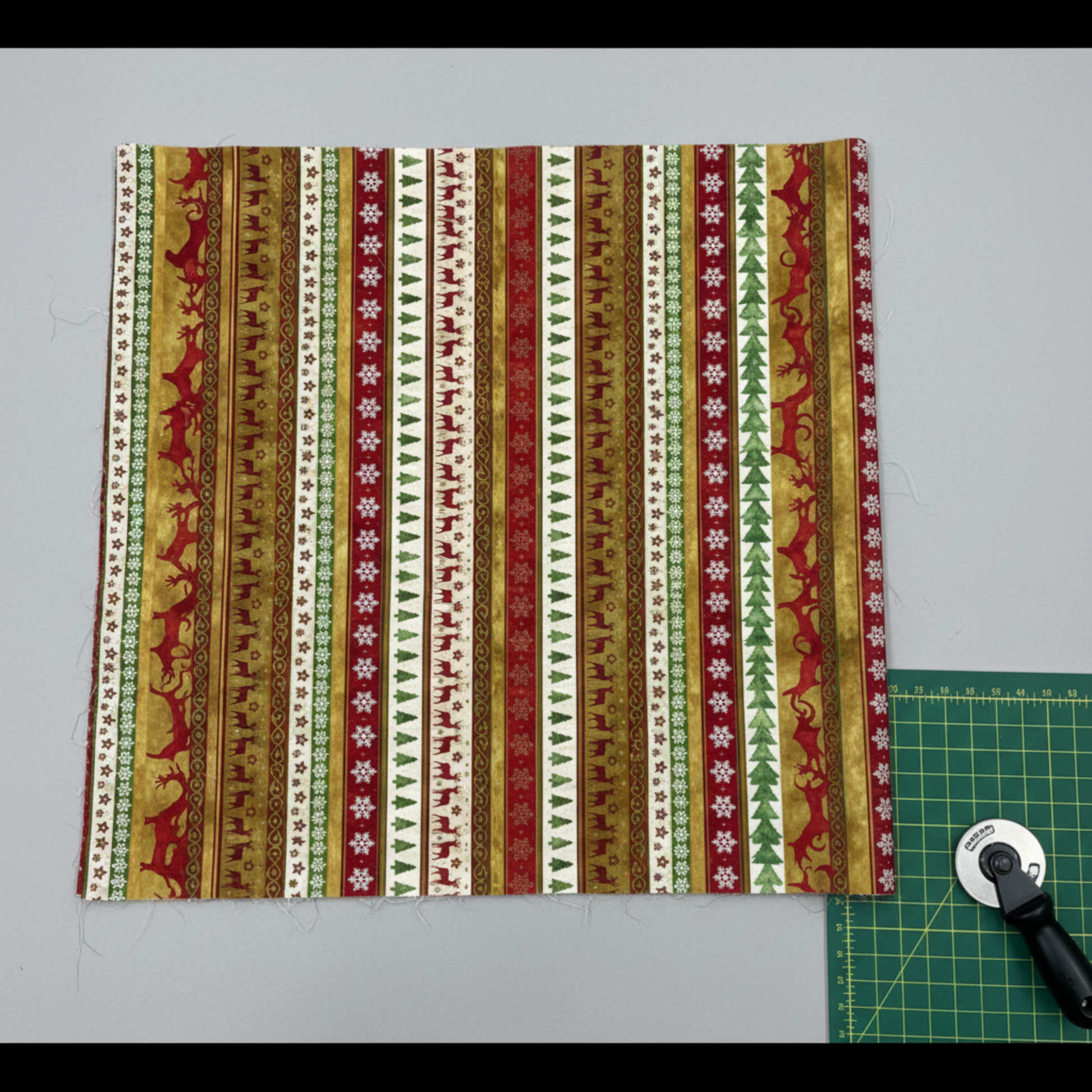 Winter Solstice II Multi Stripe Metallic quilting cotton by Jason Yenter for In The Beginning Fabrics — fabric flat lay showing full Christmas stripe repeat with snowflakes, trees, reindeer, and stars in red, cream, gold, and white with metallic accents, shown with cutting mat and rotary cutter. Quilt blocks shown for inspiration. Coordinating fabrics and quilt patterns are available at IraRobi Quilts.