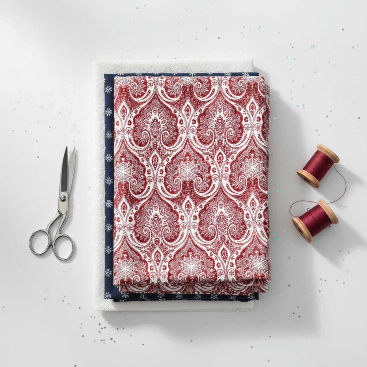 Nature's Winter Red Snowflake Ogee quilting cotton by Jason Yenter for In The Beginning Fabrics — folded as hero on a coordinating fabric stack with silver scissors and deep red thread spools on a white surface. Coordinating fabrics and quilt patterns available at IraRobi Quilts.