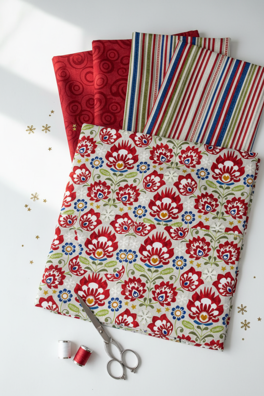 NYSNO New Snow Multicolor Floral quilting cotton by Jason Yenter for In The Beginning Fabrics — vertical flat lay as hero fabric fanned over coordinating fabrics with silver scissors and thread spools, gold snowflake and star confetti on a white surface. Coordinating fabrics and quilt patterns available at IraRobi Quilts.